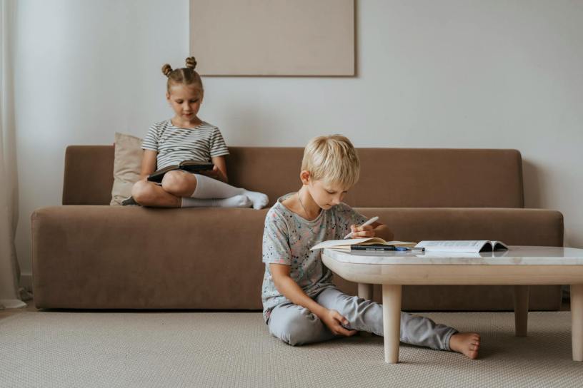A boy studies while a girl relaxes on the sofa, both in a comfortable indoor room setting.